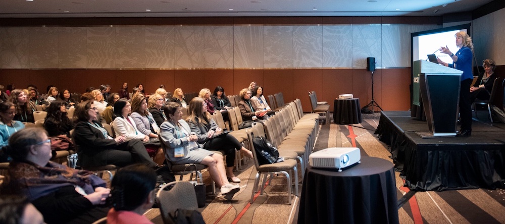 A scene in a conference room where many professional women are listening to another woman on the stage delivering a presentation