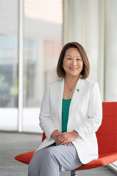 AACSB CEO Lily Bi sits with hands folded in her lap as she sits in a modern red chair with brightly lit tall windows and white curtains in the background. She has shoulder-length side-parted dark brown hair and bright smile and wears a delicate necklace and a white blazer over an emerald green shirt with an AACSB green-and-blue logo pin on its lapel and liight gray pants.
