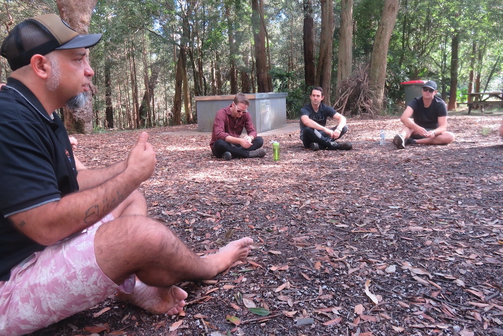 In an outdoor setting, an Aboriginal elder asks business students to speak and listens carefully to each observation before offering his own thoughts.