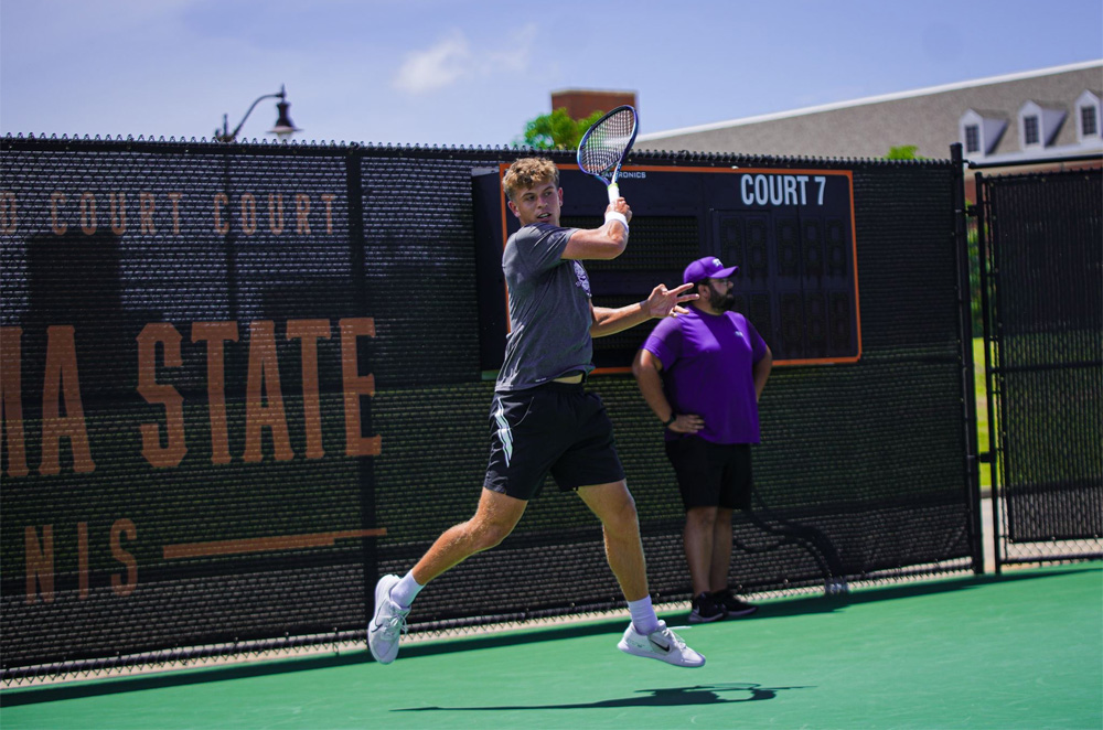 Author Luke Swan in a blue polo shirt and dark blue shorts, shown on the court with his racket during a tennis match, in mid-air with one leg behind him, as he follows through on his swing after hitting the ball. A black scoreboard wall and line judge wearing black shorts and a purple shirt are behind him.