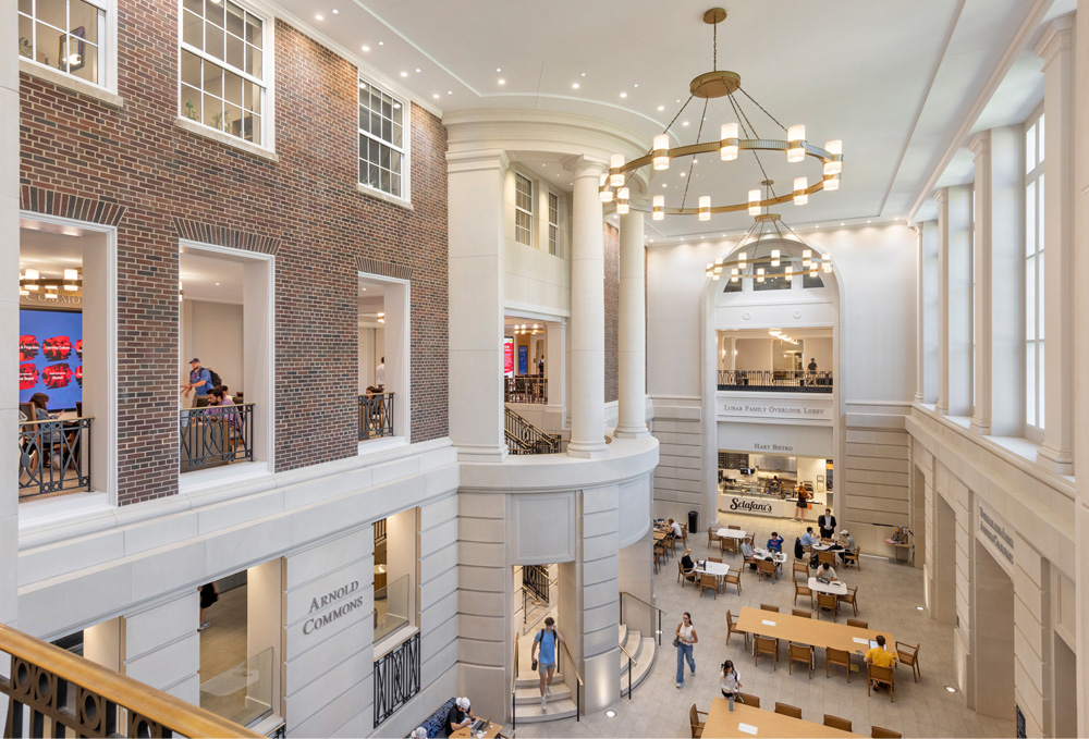 Arnold Commons at Southern Methodist University's Cox School of Business, a spacious bright commons with tall ceilings, brick and white architectural details, large white columns, cream-colored tile floors, and modern circle chandeliers, where students sit at well-spaced tables studying, talking, and using laptops.